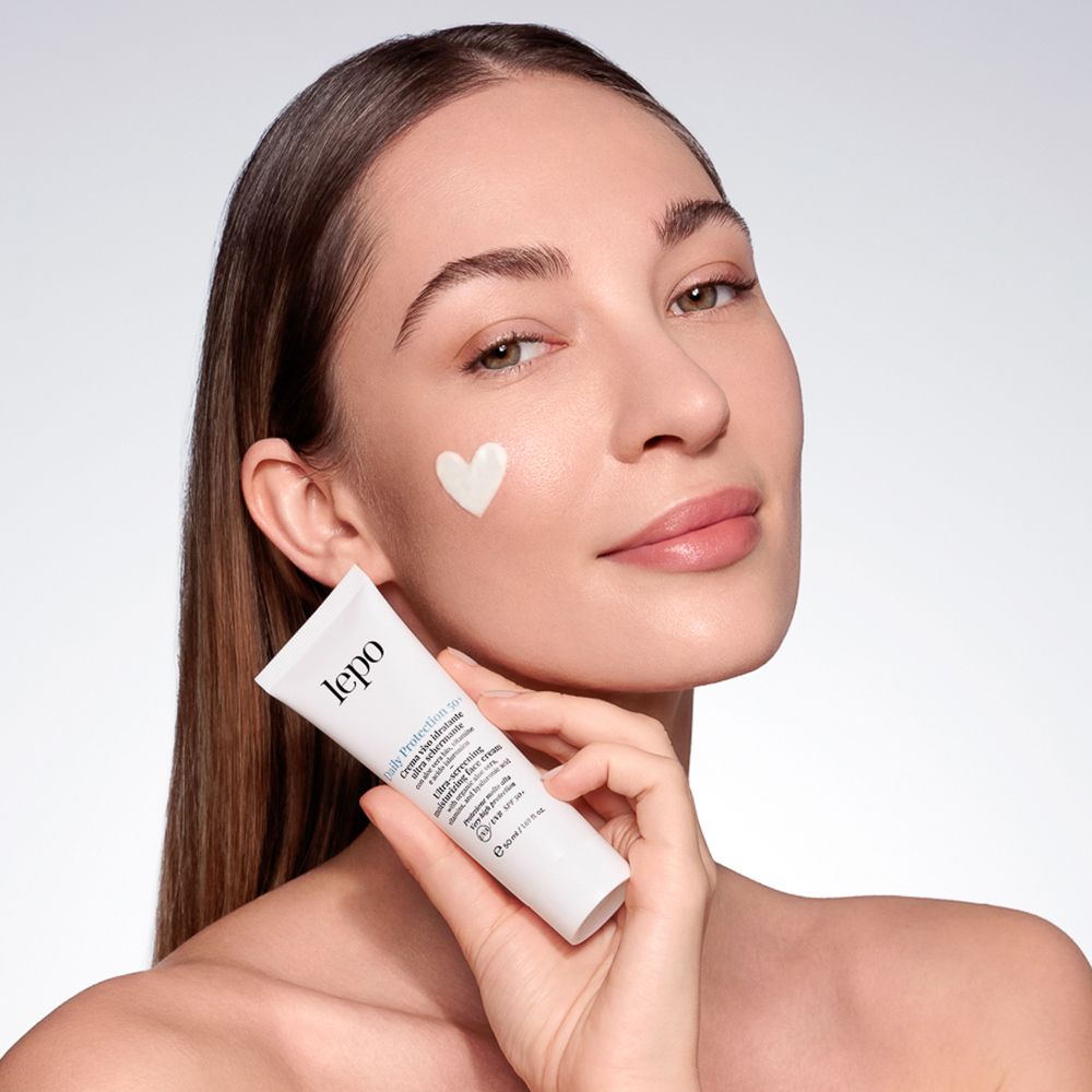 Donna con crema sulla guancia. Tubo in mano. Testo: Daily Protection 50+, crema viso ultra schermante. Marchio: lepo.