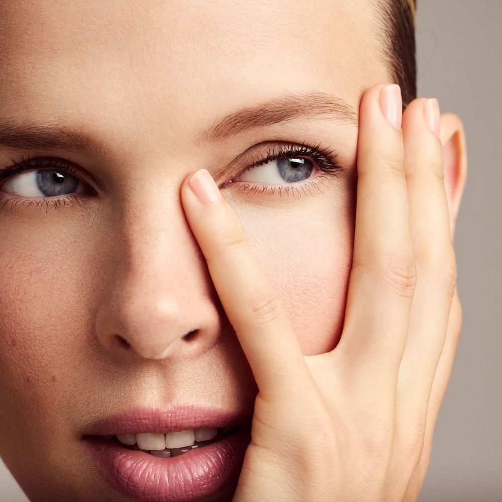 Primo piano di un viso. Una mano copre l'occhio. Pelle chiara, occhi blu, labbra rosa.