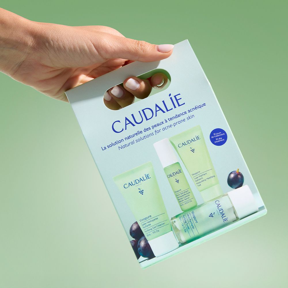 Mano che tiene un set Caudalie. Confezione verde con gel detergente, lozione, siero e idratante. Logo e testo.