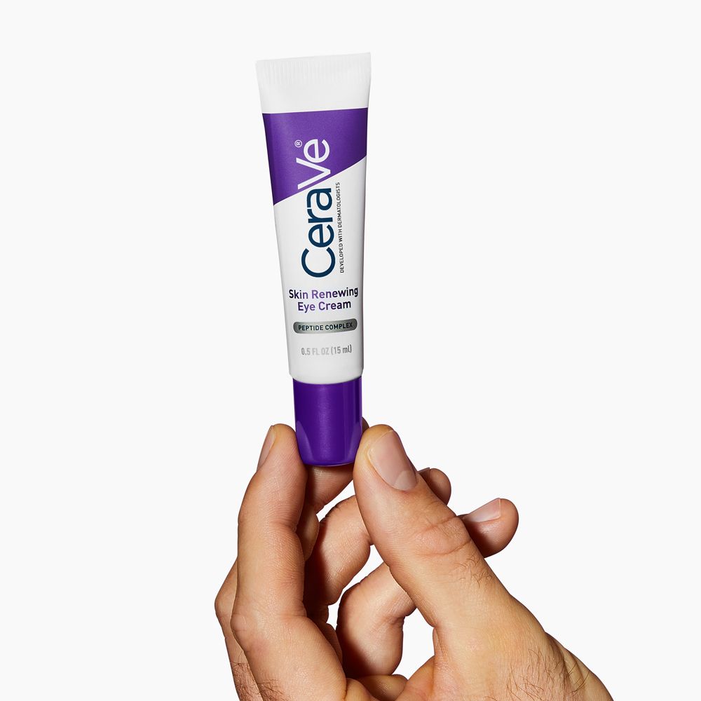 Mano che tiene il tubo di crema contorno occhi CeraVe Skin Renewing. Bianco, viola. Prodotto visibile. Ambiente luminoso.