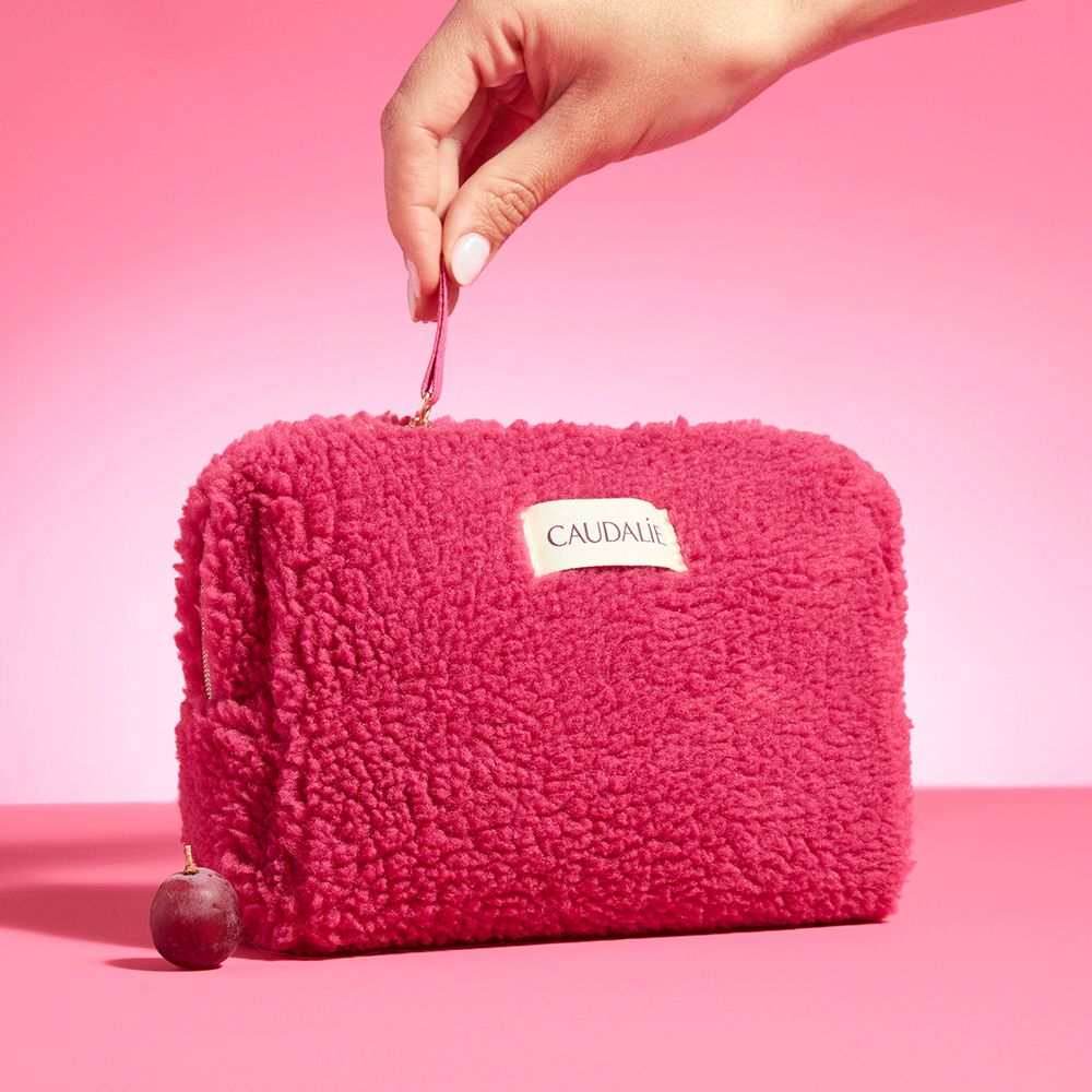 Trousse rosa con logo Caudalie. Mano tiene la zip. Uva accanto.