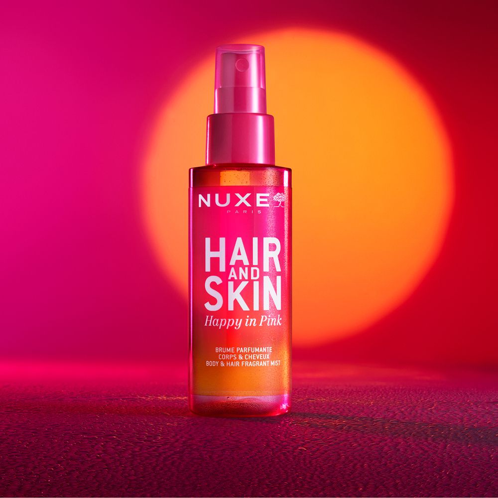 Flacone rosa con vaporizzatore. Scritta: Nuxe, Hair and Skin, Happy in Pink. Sfondo rosso e arancione.