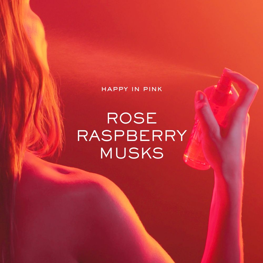 Mano che spruzza una nebbia rosa da un flacone. Scritta: Happy in Pink, Rose Raspberry Musks. Sfondo rosso.