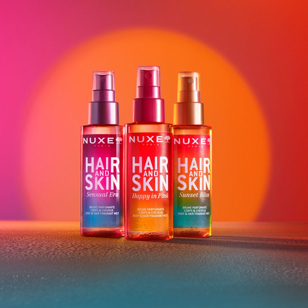 Tre flaconi con vaporizzatori. Scritta: Nuxe, Hair and Skin. Diversi profumi. Sfondo: rosso, arancione, blu.