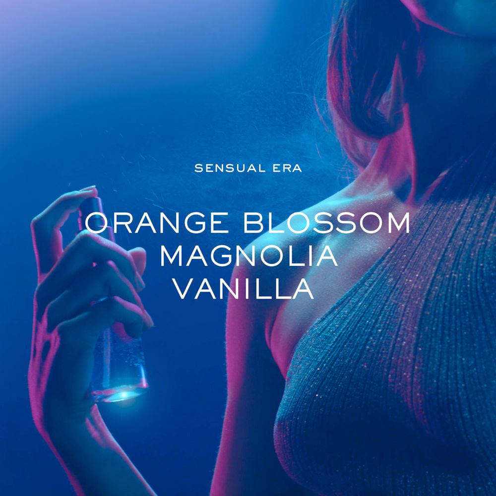 Primo piano di una donna che tiene un flacone spray. Testo: Sensual Era, Fiori d'arancio, Magnolia, Vaniglia.