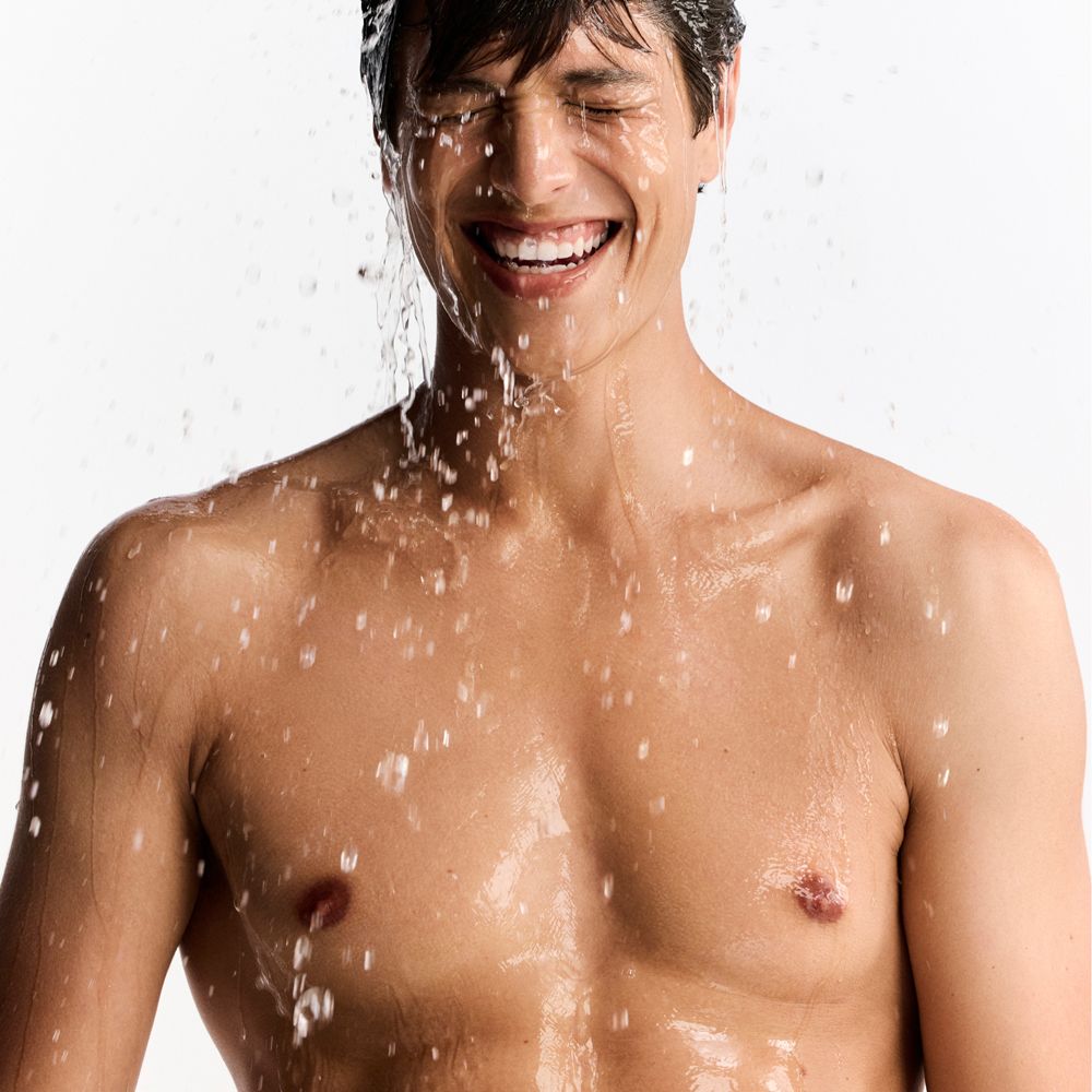 Uomo con acqua sul viso. Sorride. L'acqua schizza sul suo viso e sul busto. Ambiente luminoso.