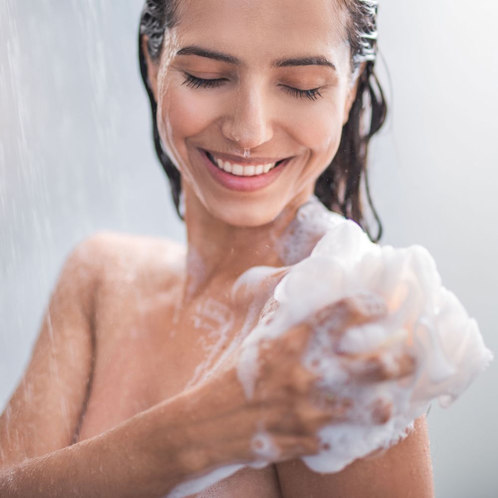 Donna che fa la doccia e si lava con una spugna. Sorride. Sfondo sfocato.