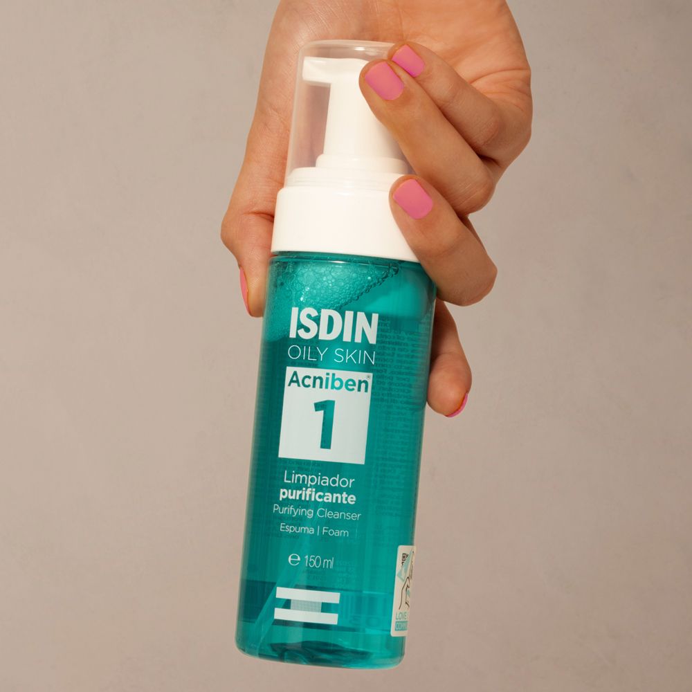 Mano che tiene un flacone. Scritta: ISDIN, OILY SKIN, Acniben 1. Unghie smaltate di rosa.