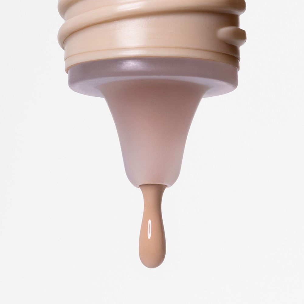 Primo piano di un'apertura a goccia. Una goccia di fondotinta color pelle. Il collo del flacone è beige.