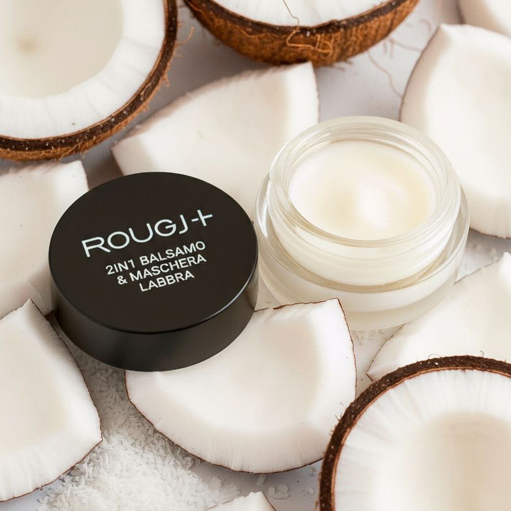 Vasetto aperto con balsamo, circondato da pezzi di cocco. Coperchio nero con testo bianco: Rougj+, 2in1 Balsamo & Maschera Labbra.