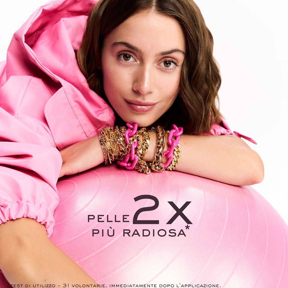Donna appoggiata su palla rosa. Abbigliamento rosa e gioielli. Testo: Pelle Più Radiosa 2X.