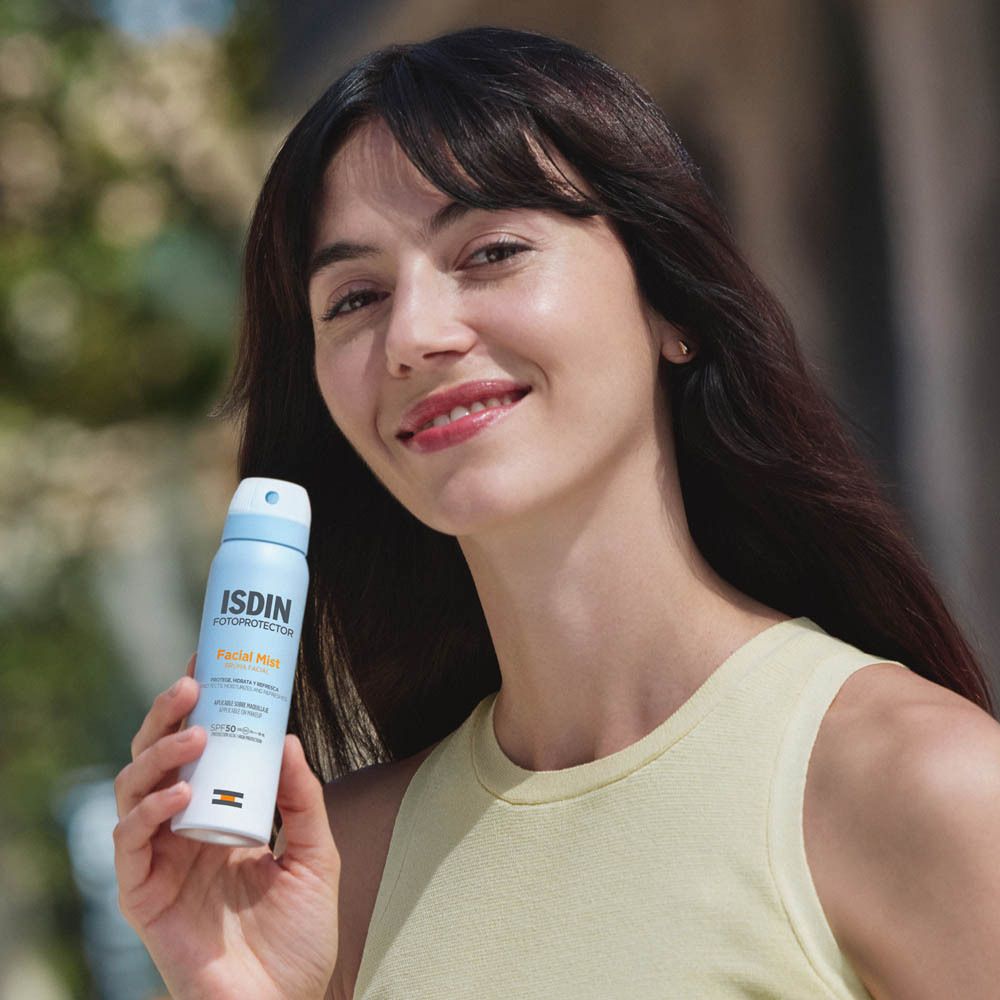 Donna tiene ISDIN Fotoprotector Bruma Viso SPF 50. Flacone spray blu. La donna sorride. Sfondo sfocato.
