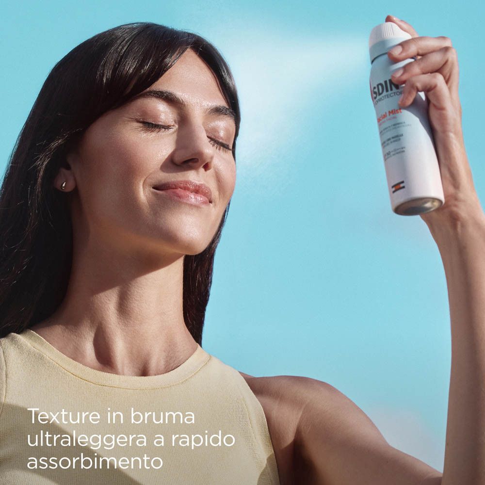 Donna spruzza ISDIN Fotoprotector Bruma Viso SPF 50 sul viso. Sfondo blu. Nome del prodotto visibile.