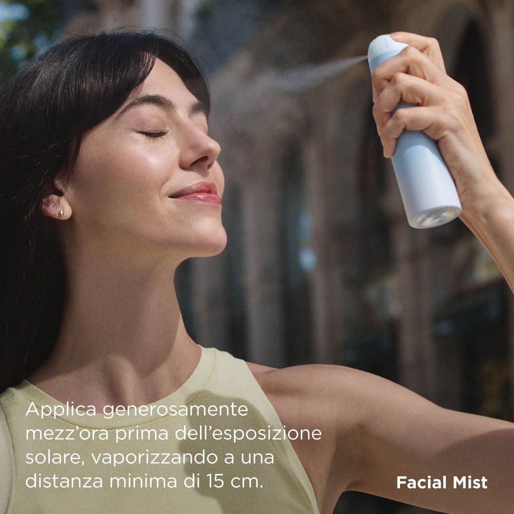 Donna spruzza ISDIN Fotoprotector Bruma Viso SPF 50 sul viso. Sfondo blu. Nome del prodotto visibile.