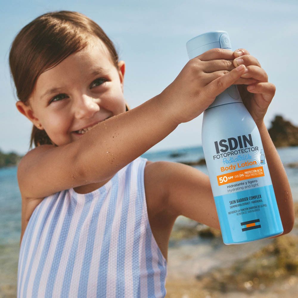 Bambina tiene in mano un flacone spray blu chiaro con logo ISDIN. Sfondo spiaggia. La bambina sorride.