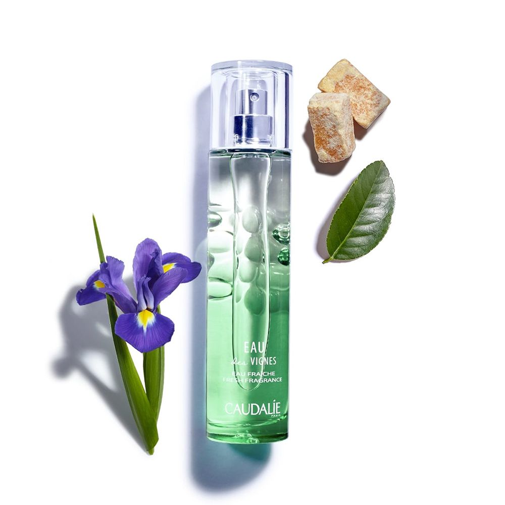 Flacone di profumo verde, accanto: fiore di iris, foglie, zollette di zucchero. Scritta: Eau des Vignes, Caudalie.