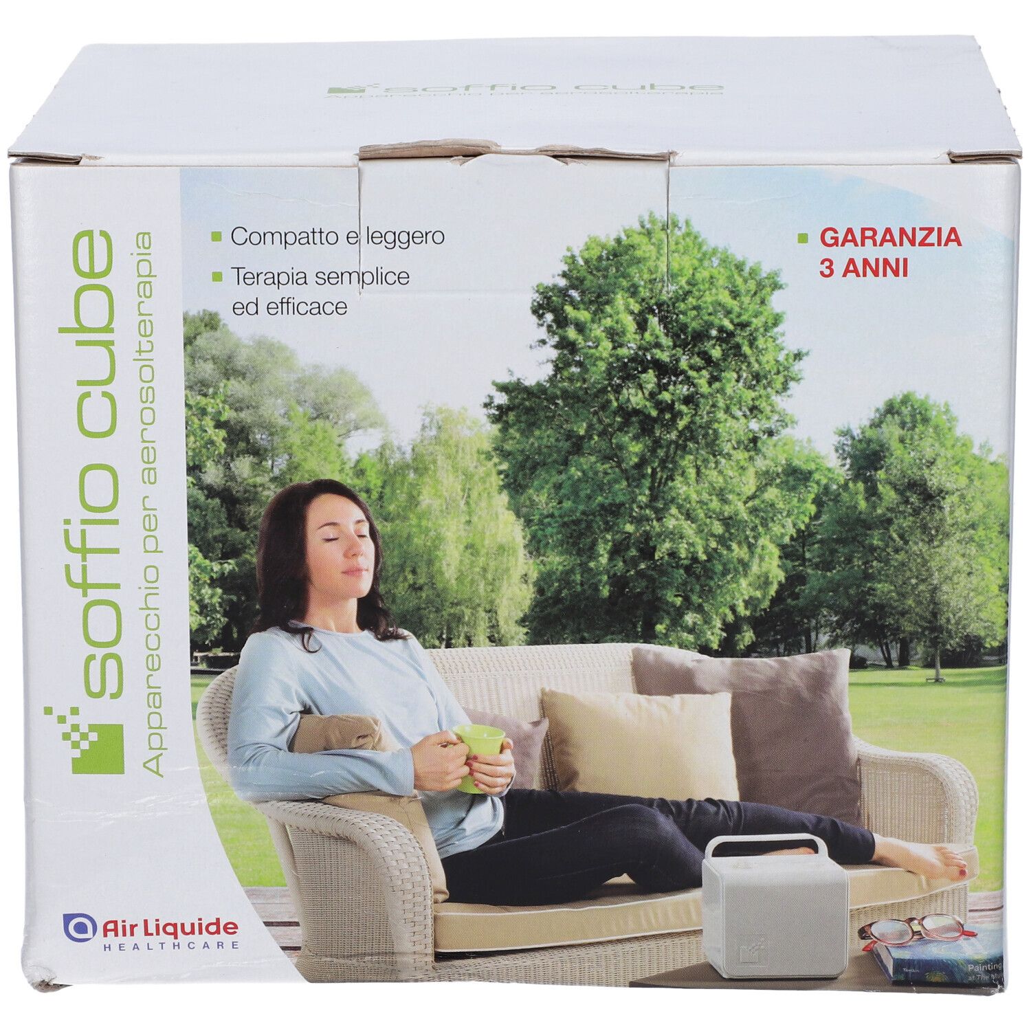 Scatola dell'apparecchio per aerosol Soffio Cube. Scritte: Garanzia 3 anni, Air Liquide Healthcare. Donna su divano con dispositivo.