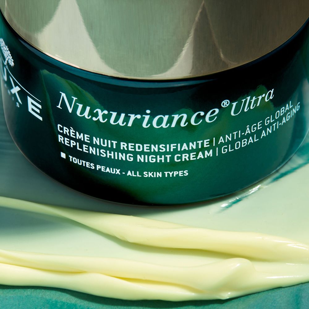 Primo piano del prodotto. Scritta: Nuxuriance Ultra, crema notte. Texture crema su superficie chiara.
