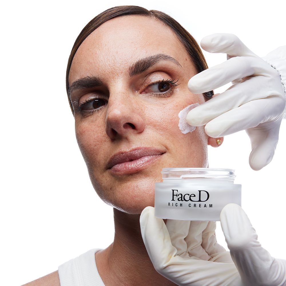 Mani con guanti applicano crema sul viso di una donna. Face D Rich Cream.