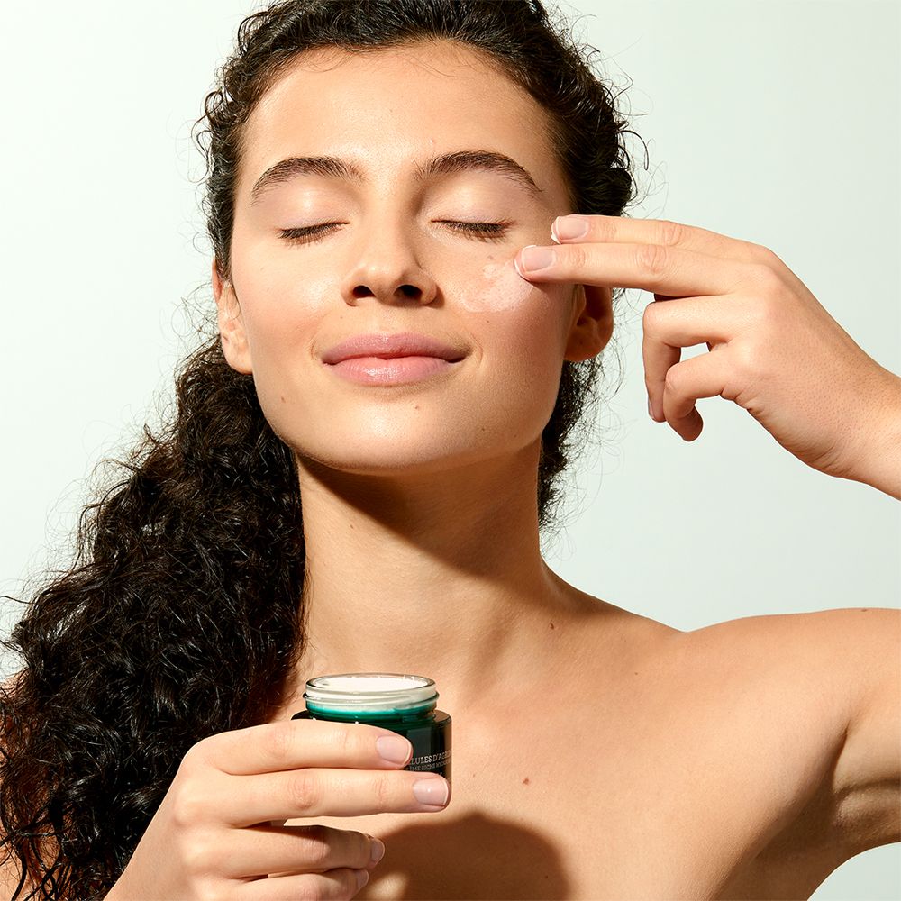 Donna con occhi chiusi, tocca il viso con un dito. Piccolo vaso di crema visibile. Nessun altro dettaglio.