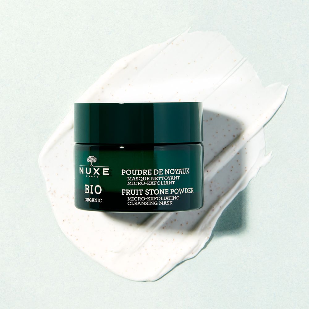 Vasetto verde scuro con coperchio e prodotto su sfondo bianco. Scritta: NUXE BIO, Poudre de Noyaux, Fruit Stone Powder.