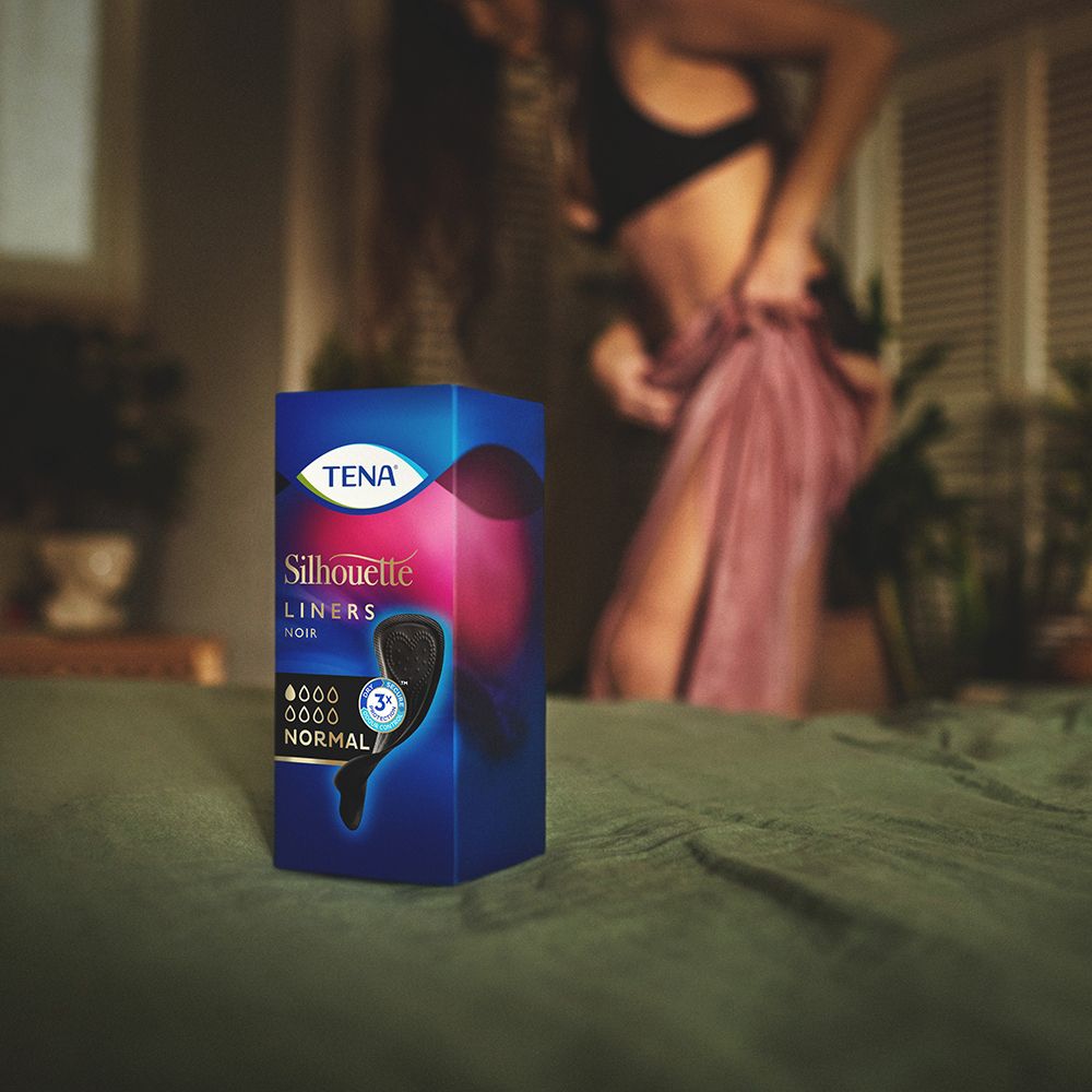 Confezione del prodotto su un letto. Marca TENA, Silhouette Noir, Normal. Sfondo: donna che si veste.