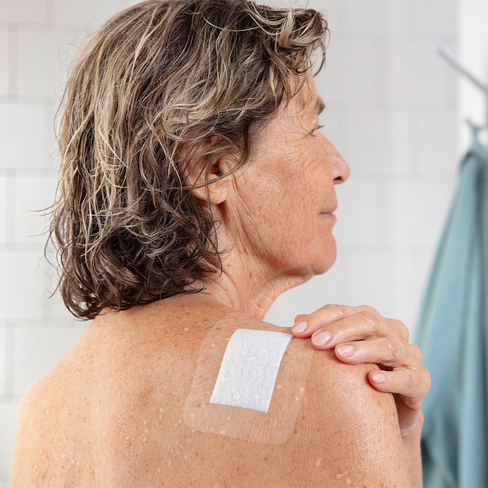Donna con cerotto sulla spalla. Cerotto trasparente, rettangolare. Contatto con la pelle, doccia sullo sfondo.