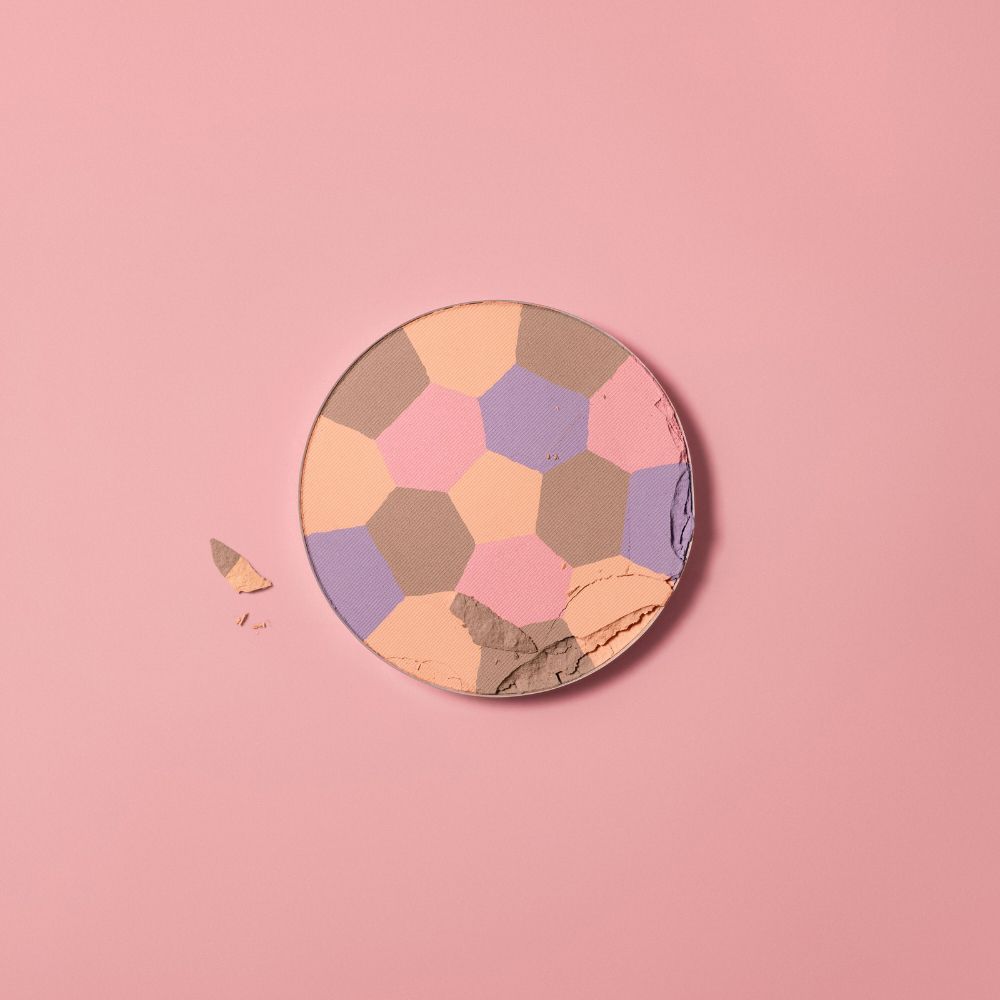 Cipria rotonda con campi di colore esagonali. Un pezzo rotto accanto. Sfondo rosa.