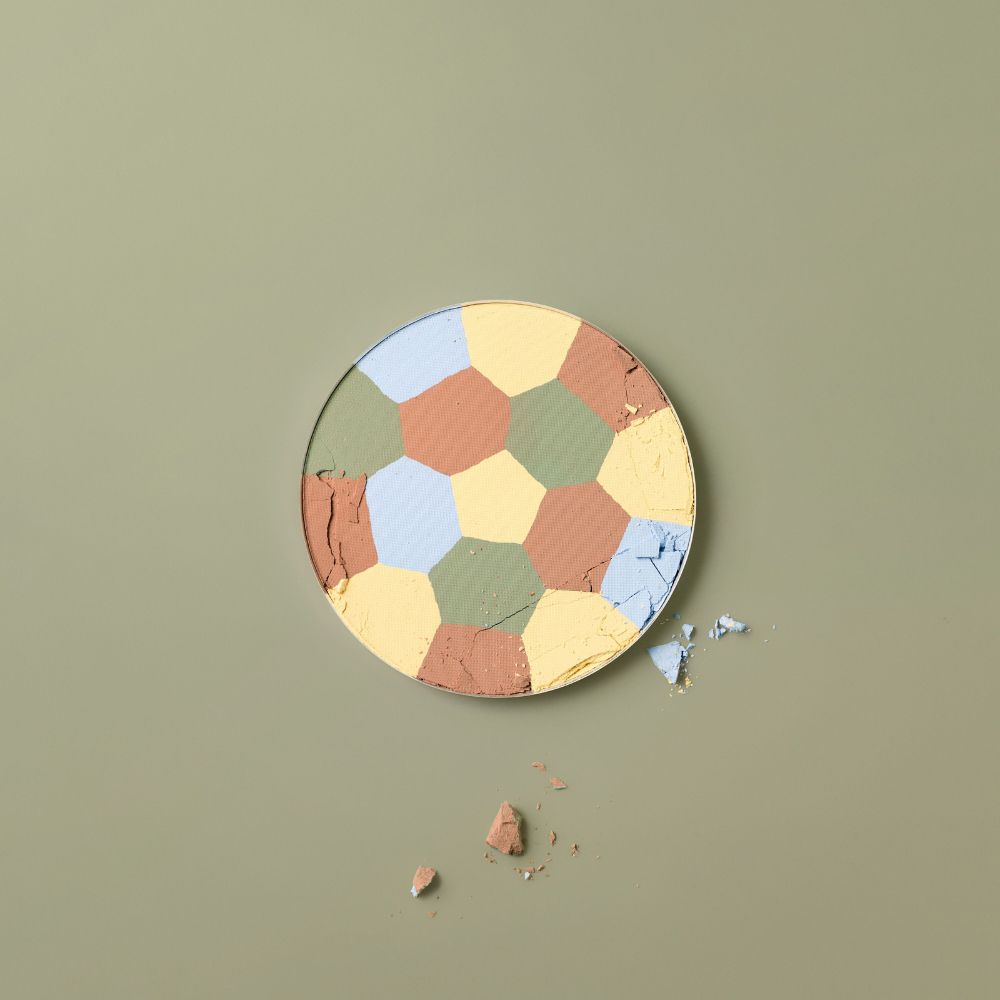 Cipria compatta rotonda con segmenti colorati, parzialmente rotta, su sfondo verde.