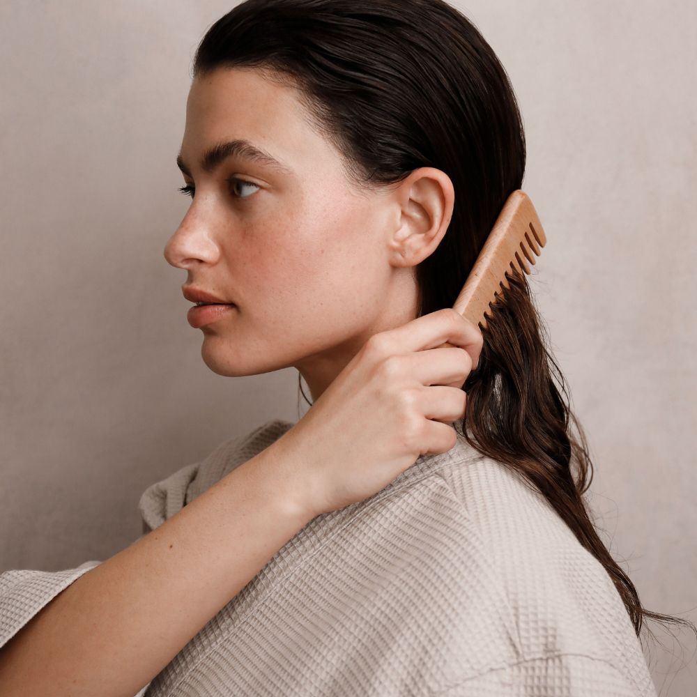 Donna che si pettina i capelli bagnati con un pettine di legno. Indossa un top beige.