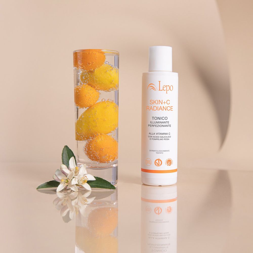 Flacone bianco LEPO Skin+C Radiance Tonico accanto a bicchiere con agrumi. Testo: Skin+C Radiance, Tonico.