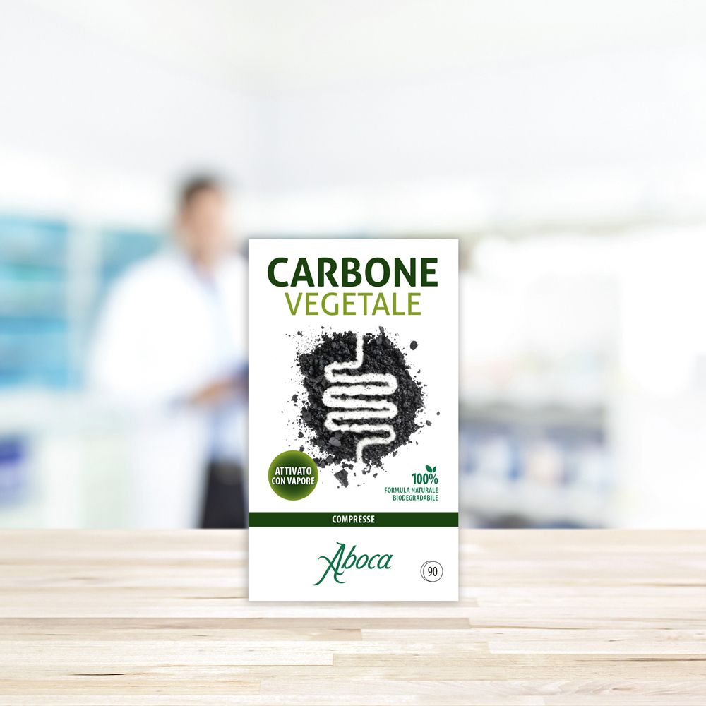 Confezione prodotto su tavolo in legno. Scatola Carbone Vegetale. Logo Aboca. Sfondo: farmacia sfocata.