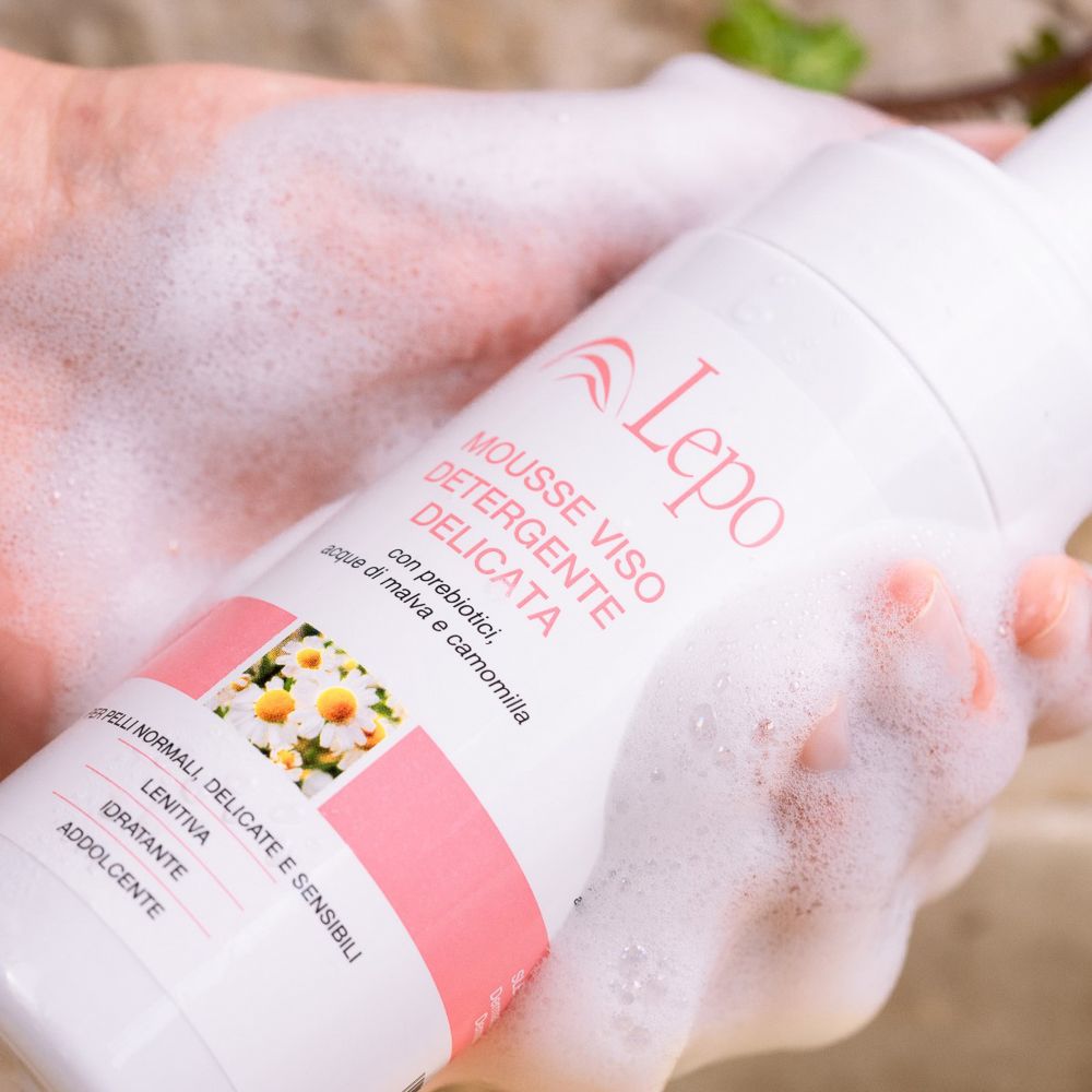Mani che tengono un flacone. Schiuma sulla mano. Flacone con etichetta rosa. Testo: MOUSSE VISO DETERGENTE DELICATA.