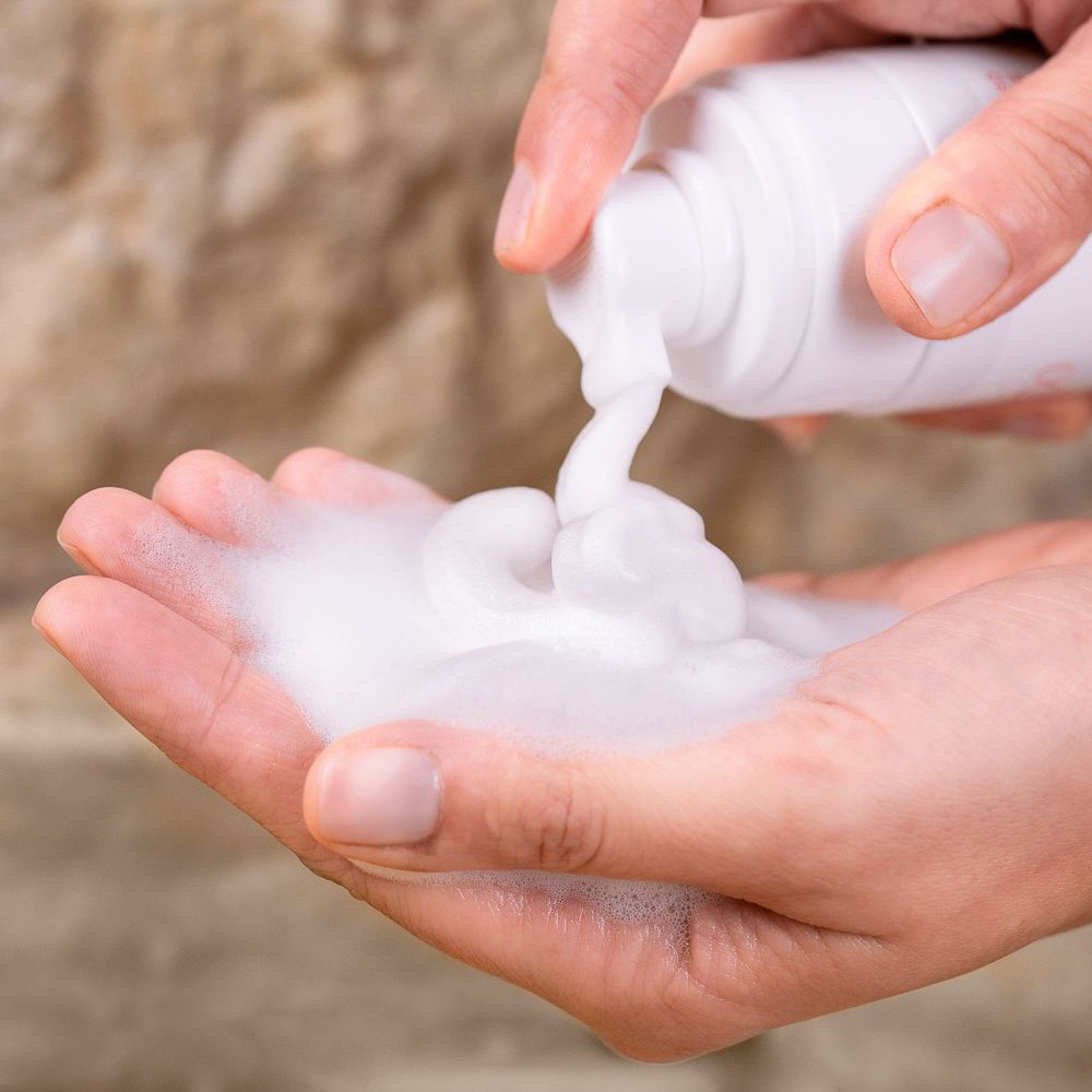 Mani che tengono schiuma bianca. Il prodotto viene erogato da un flacone bianco.