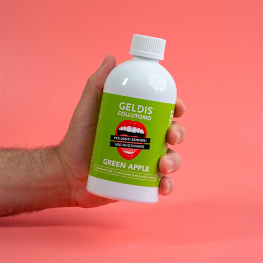 Mano che tiene una bottiglia bianca con etichetta verde. Scritta GELDIS Collutorio, Green Apple. Sfondo rosa.