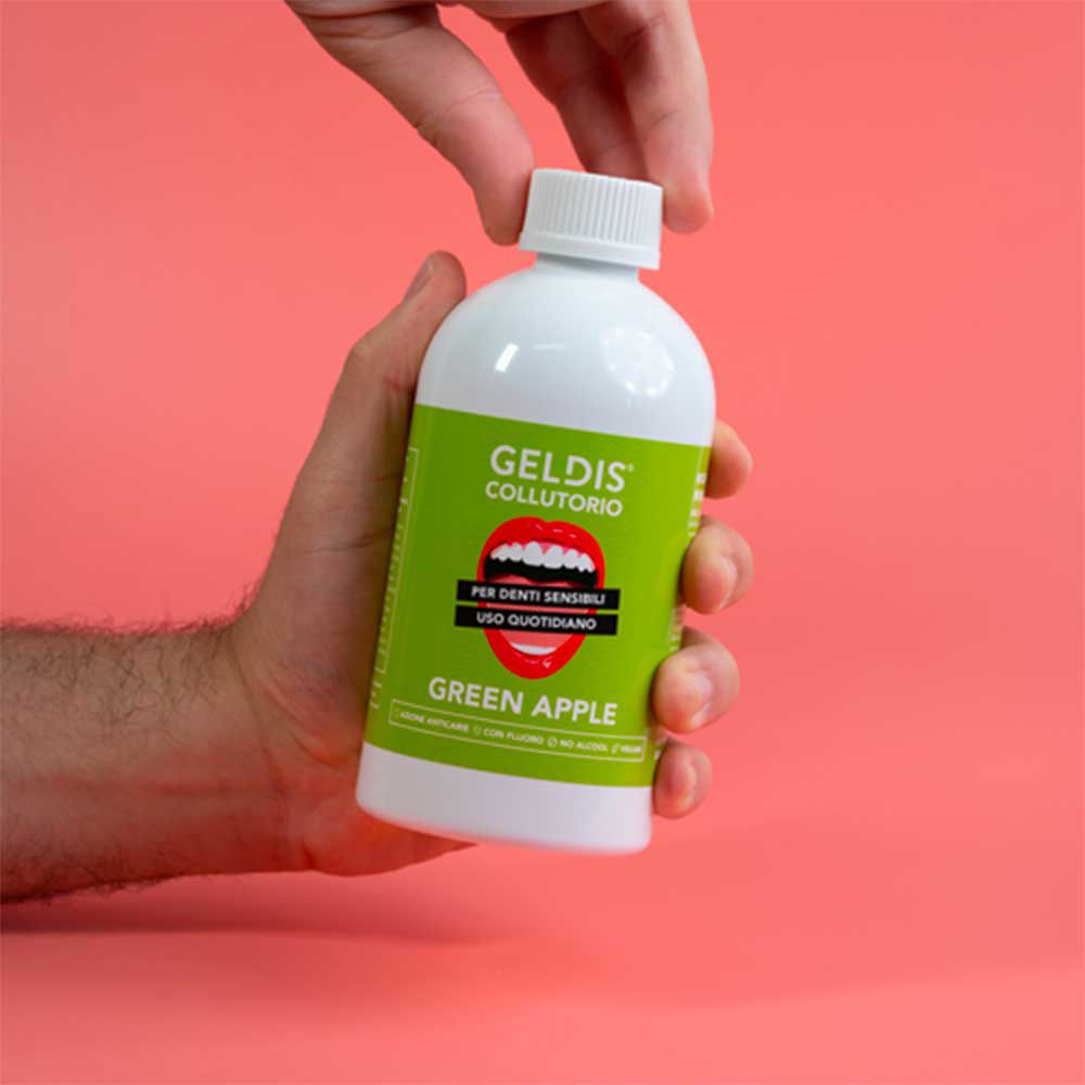 Mano che apre una bottiglia bianca con etichetta verde. Scritta GELDIS Collutorio, Green Apple. Sfondo rosa.