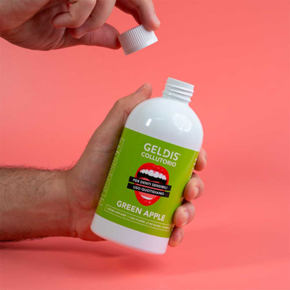 Mano che tiene una bottiglia bianca aperta con etichetta verde. Scritta GELDIS Collutorio, Green Apple. Sfondo rosa.