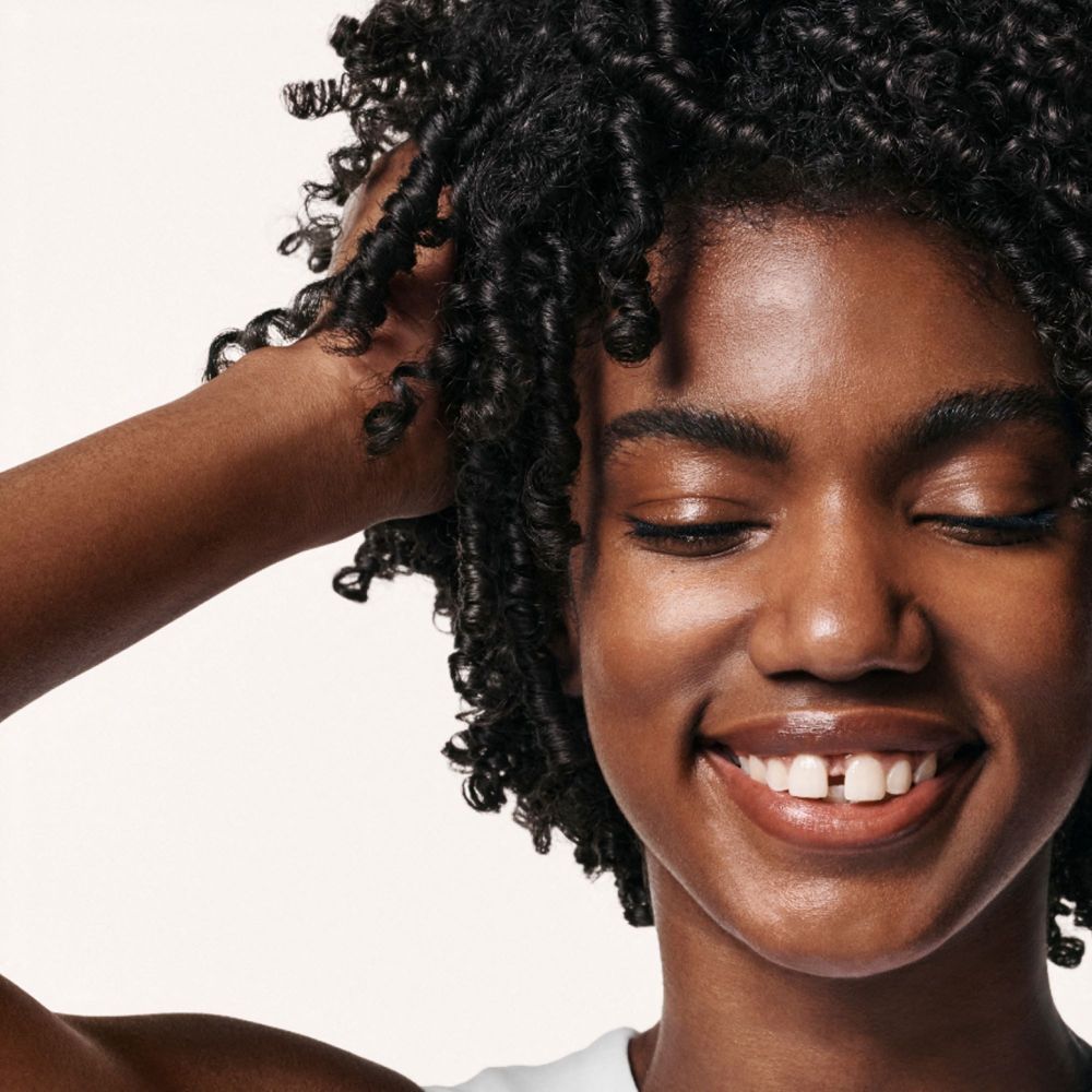 Donna con capelli ricci. Occhi chiusi, sorridente. Mani tra i capelli. Pelle chiara. Probabilmente cura dei capelli.