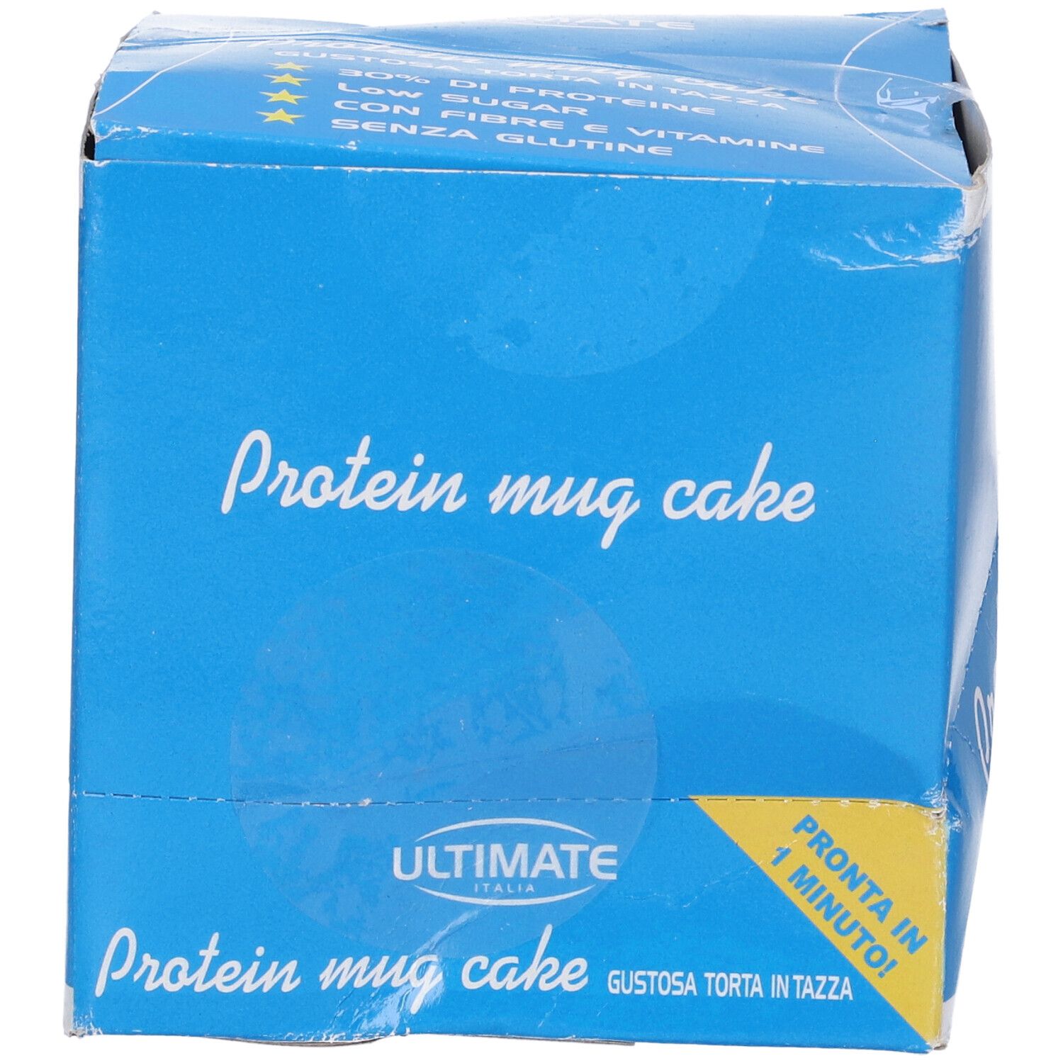 Scatola blu con scritta "Protein mug cake". Contiene informazioni su proteine, zuccheri, fibre e vitamine. Etichetta gialla: "Pronta in 1 minuto!"