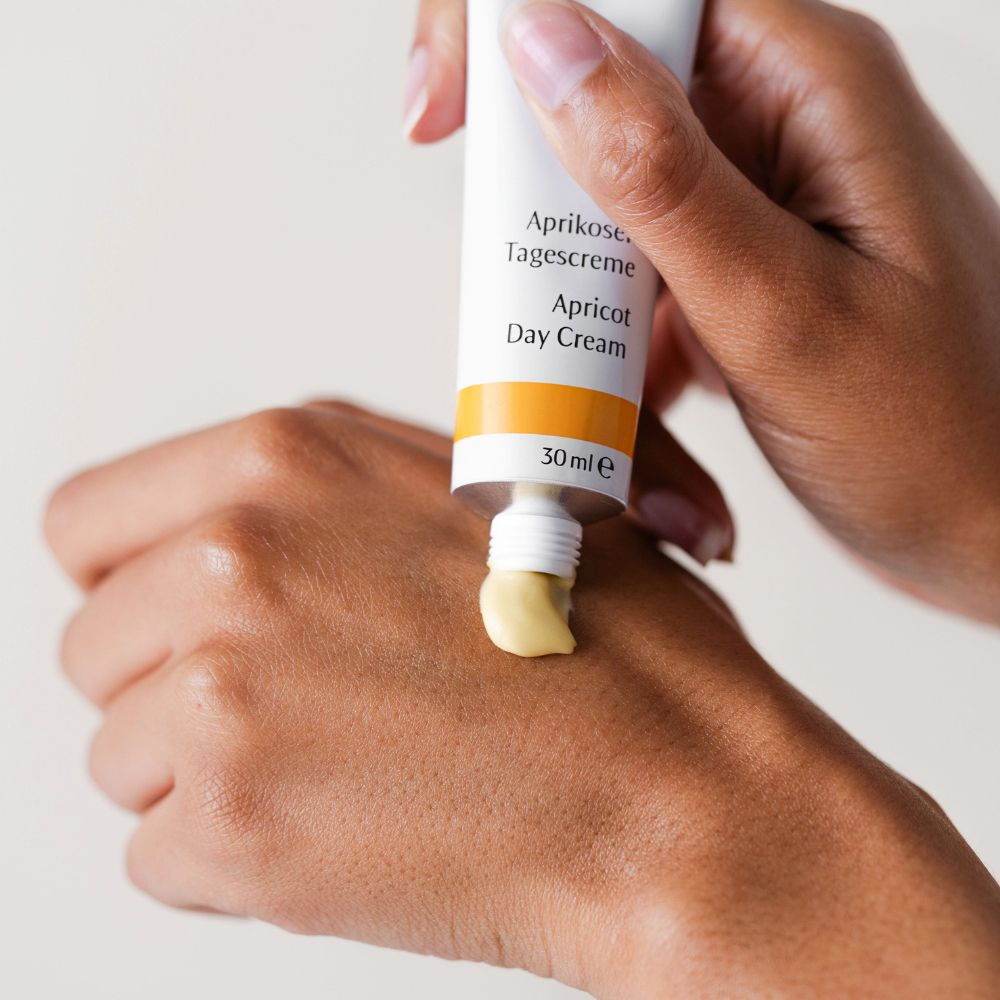 Mano che preme la crema dal tubetto. Scritta: Aprikosen Tagescreme, Apricot Day Cream, 30ml e. Crema giallastra.