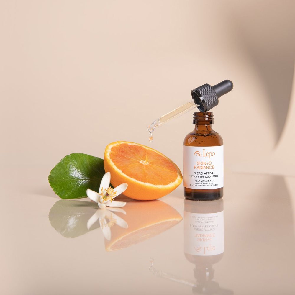 Prodotto per la cura della pelle con flacone, arancia e fiore. Gocce cadono dalla pipetta. Scritta: Skin+C Radiance Siero Attivo.