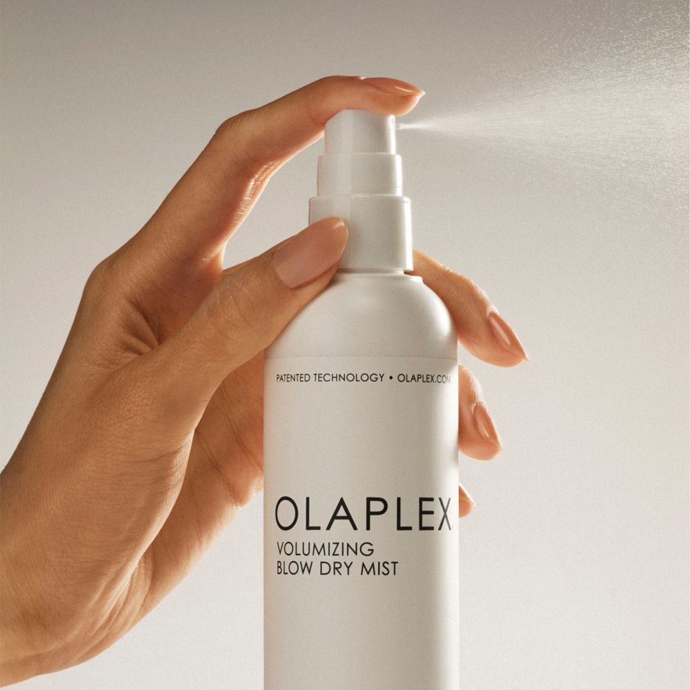 Mano che spruzza Olaplex Volumizing Blow Dry Mist. Flacone bianco, spruzzo visibile.