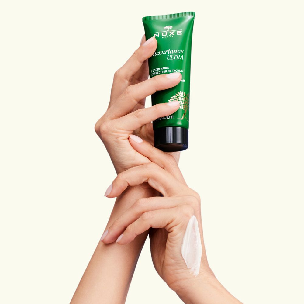 Mani che tengono un tubo verde Nuxe Nuxuriance Ultra. Crema su una mano. Testo: Le soin mains correcteur de taches, The dark spot correcting hand cream.