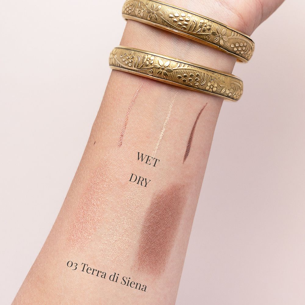 Braccio con tre campioni di ombretto. Testo: Wet, Dry, 03 Terra di Siena. Bracciali dorati.