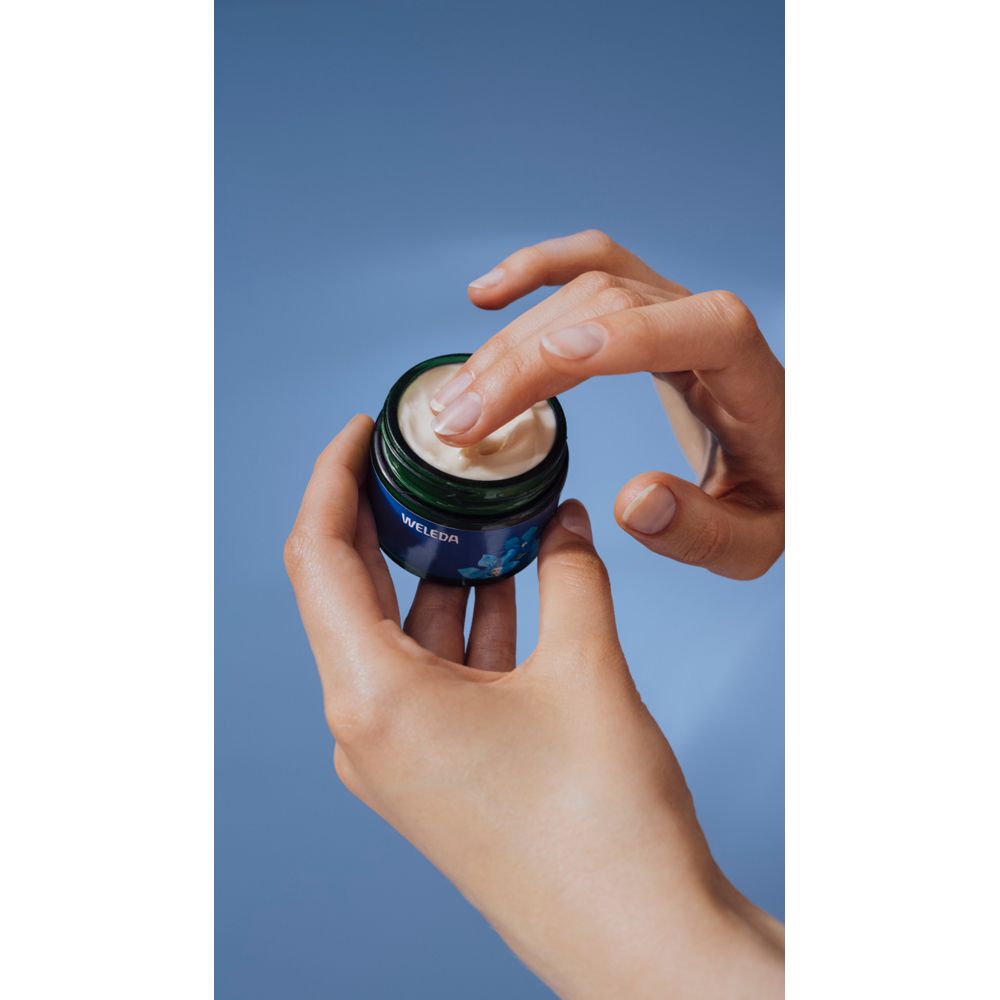 Mani che tengono un vasetto di crema aperto. Scritta: WELEDA, Crema Notte Contouring. Sfondo blu.