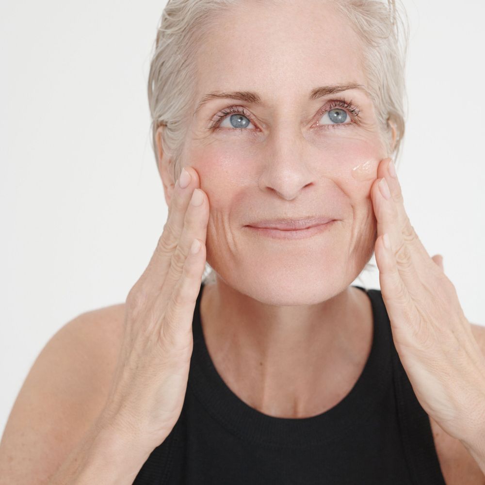 Donna anziana applica siero sul viso. Top nero. Nessuna denominazione del prodotto visibile.