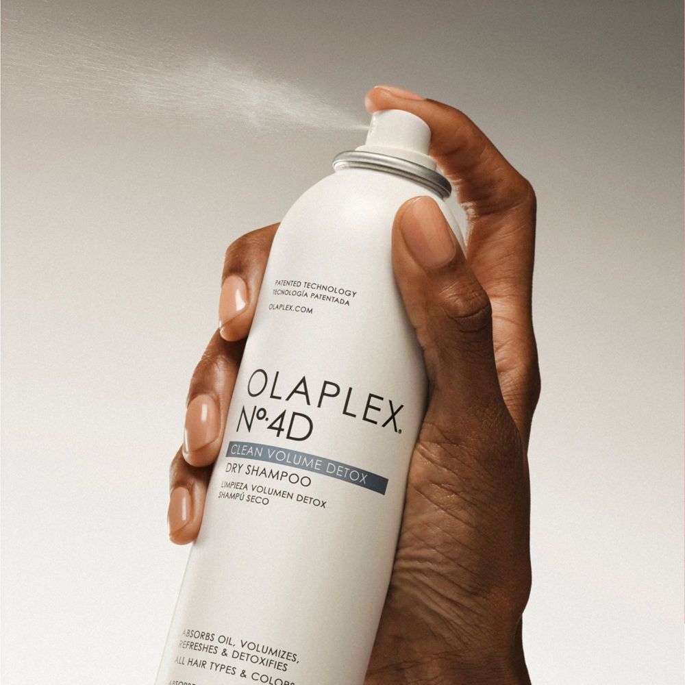 Mano che tiene lo spray shampoo secco Olaplex N°.4D. Nebbia spray visibile. Nome e dettagli del prodotto stampati.