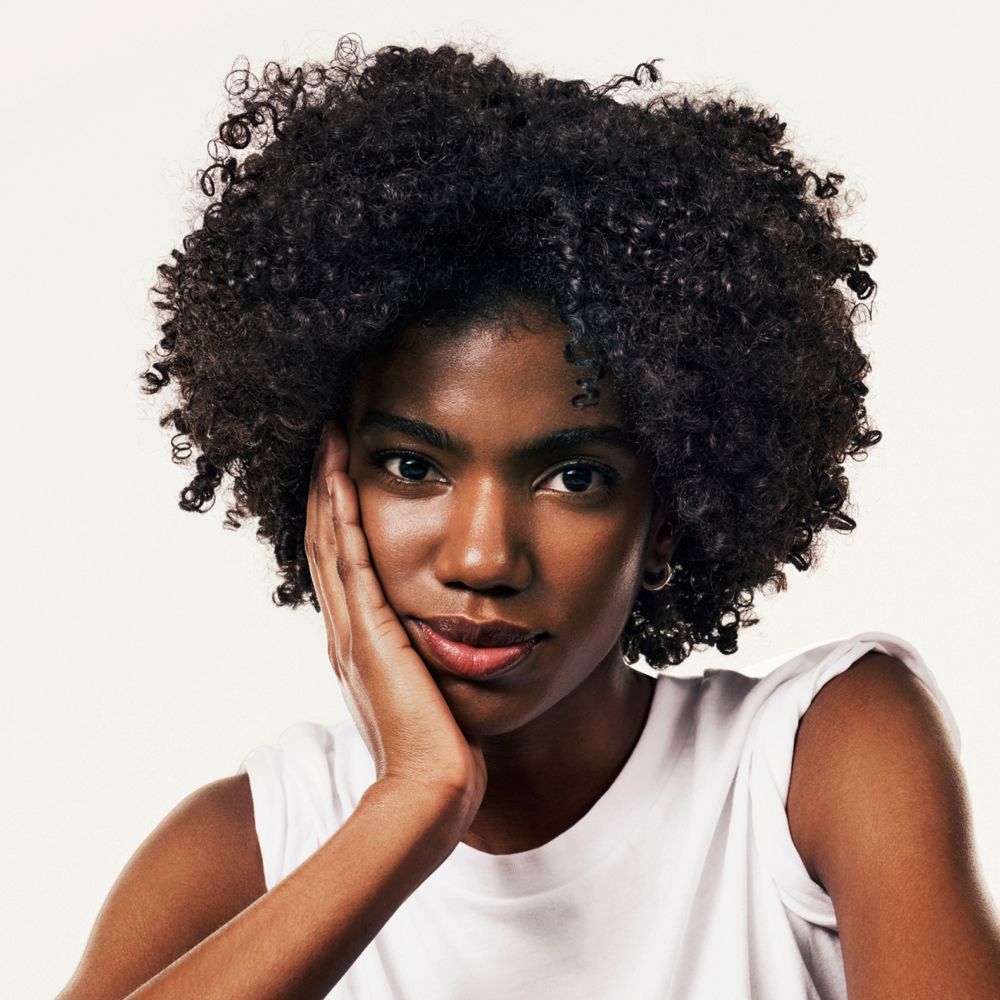 Ritratto di donna con capelli ricci. Si tocca la guancia. Canotta bianca.