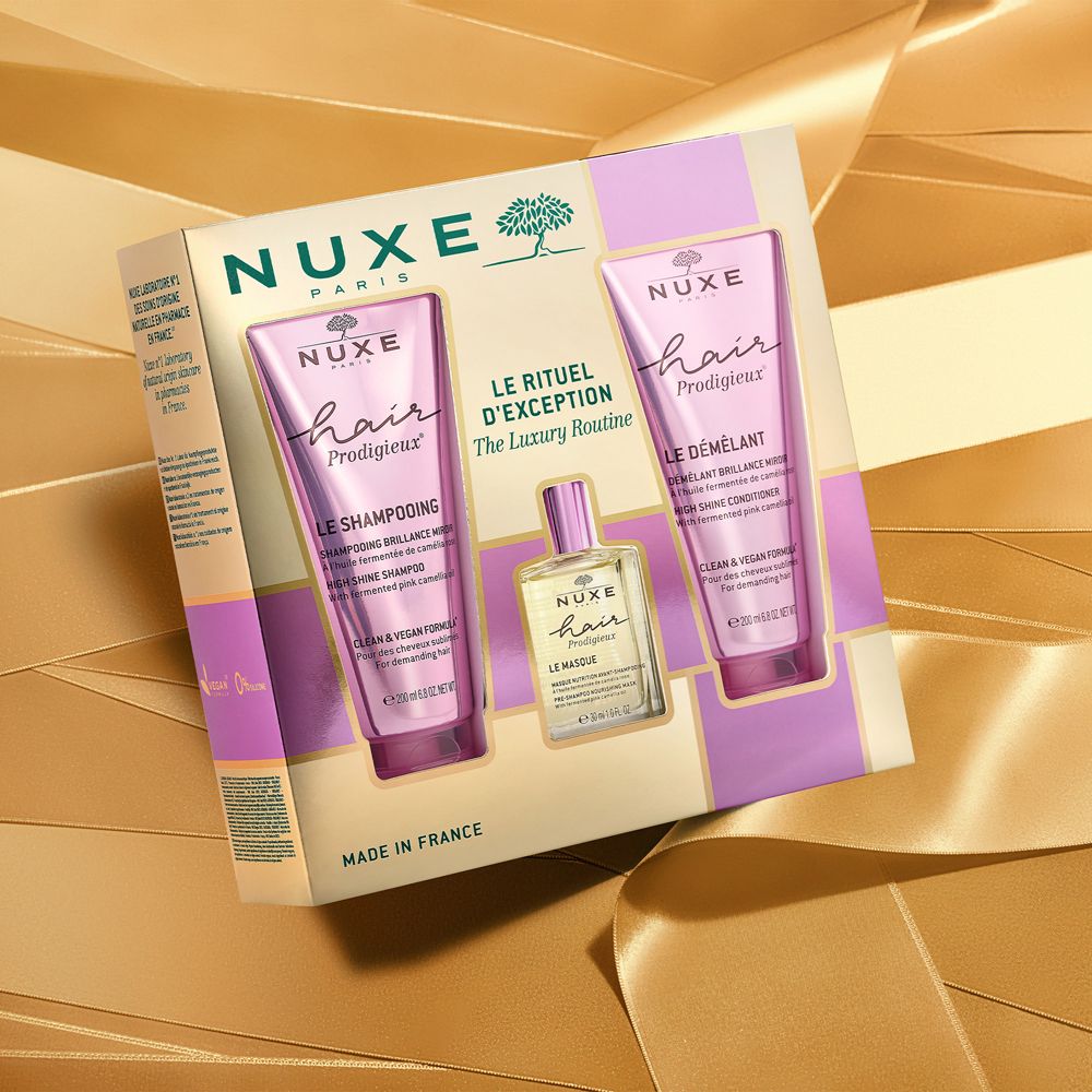 Confezione regalo Nuxe Hair Prodigieux. Due tubi e una bottiglia. Fiocco dorato sullo sfondo. Confezione oro e viola.