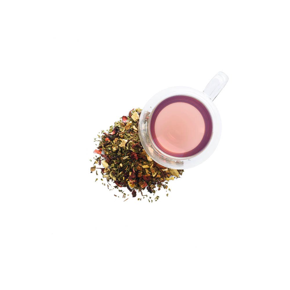 Tazza di vetro con tè alle erbe e tè sfuso accanto. Il tè è rosa. Le erbe sfuse sono verdi e rosse.